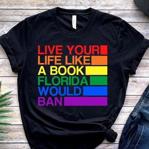 May include: Black t-shirt with the text "Live your life like a book Florida would ban" printed in rainbow colors.