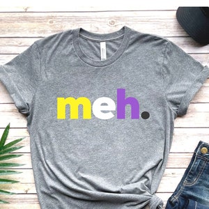 May include: Gray t-shirt with the word "meh" printed in yellow, purple, and white letters. The shirt is on a wooden background with a pair of jeans, a hat, and a plant.