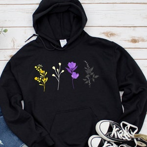 以下が含まれることがあります： 黄色、紫、白の花柄が施された黒いフード付きスウェットシャツ。花はスウェットシャツの胸に一列に並んでいます。