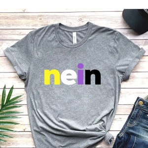 May include: A grey t-shirt with the word "nein" printed in yellow, white, and purple letters. The shirt is on a wooden surface with a green plant, blue jeans, and a black baseball cap.