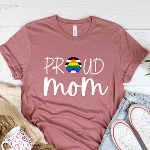 May include: Heather mauve t-shirt with a white print that says "PROUD MOM" with a rainbow pride flag in a circle in the middle of the word "PROUD".