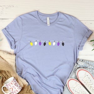 May include: A light blue t-shirt with a string of lights in the colors of the non-binary pride flag. The lights are yellow, white, purple, and black.