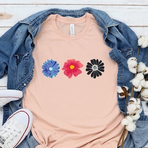 May include: A peach-colored t-shirt with three watercolor flowers printed on the front. The flowers are blue, pink, and black. The t-shirt is laid out on a white wooden surface with a denim jacket and white sneakers.