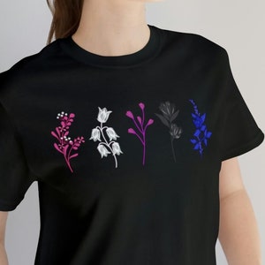 Puede incluir: Una camiseta negra con un diseño floral en rosa, blanco y azul. Las flores están estilizadas y representan la bandera del orgullo bisexual.