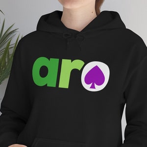 Può includere: Felpa con cappuccio nera con tasca frontale e coulisse. La parola "aro" è stampata sul petto in verde e verde chiaro, con un simbolo di picche viola all'interno di un cerchio bianco.