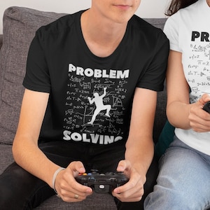 May include: Two people wearing matching t-shirts with the text "Problem Solving" and a graphic of a person climbing a wall of mathematical equations. The shirts are black and white. The people are sitting on a grey sofa and holding video game controllers.