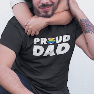 May include: A man wearing a black hat and a black t-shirt with the text "Proud Dad" and a rainbow pride symbol. He is kneeling on green grass with a child hugging him from behind.