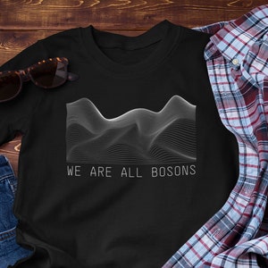 Puede incluir: Camiseta negra con un gráfico blanco de un patrón de líneas onduladas y el texto "WE ARE ALL BOSONS".