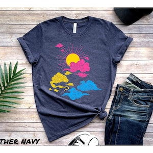 May include: A heather navy blue t-shirt with a graphic of a sun and clouds in the colors of the pansexual pride flag. The shirt is laid flat on a wooden surface.