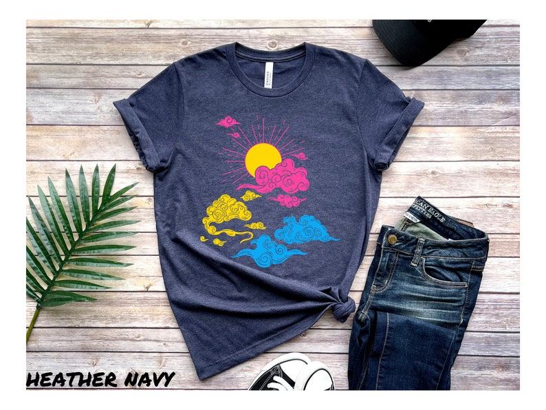 May include: A heather navy blue t-shirt with a graphic of a sun and clouds in the colors of the pansexual pride flag. The shirt is laid flat on a wooden surface.