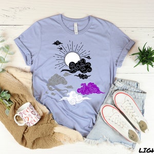 以下が含まれることがあります： 黒と白の太陽と雲のデザインが施された薄い青色のTシャツ。雲は黒、グレー、紫、白のパターンで配置されており、アセクシュアルフラッグを表しています。