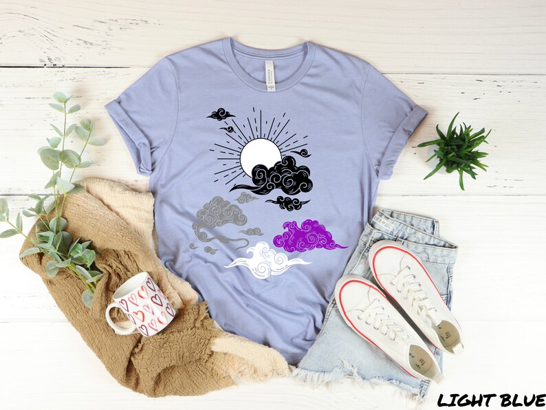 以下が含まれることがあります： 黒と白の太陽と雲のデザインが施された薄い青色のTシャツ。雲は黒、グレー、紫、白のパターンで配置されており、アセクシュアルフラッグを表しています。