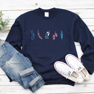 May include: A navy blue sweatshirt with a white, pink, and blue floral design on the front. The sweatshirt is paired with blue jeans and white sneakers.