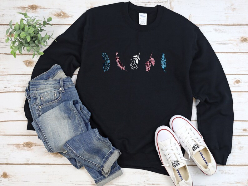 May include: Black sweatshirt with a festive design of stylized holly, berries, and pine branches in pink, blue, and green colors. The sweatshirt is laid flat on a wooden surface with a pair of white canvas shoes and blue jeans.