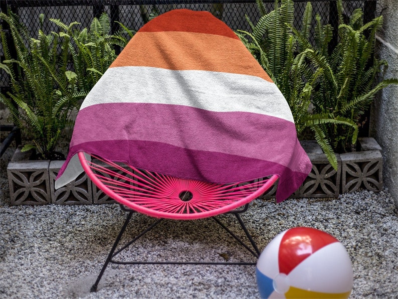 May include: A pink and black patio chair with a lesbian pride flag draped over it. A beach ball sits on the ground in front of the chair.