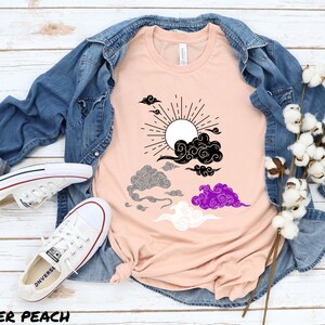 以下が含まれることがあります： ピーチ色のTシャツに、黒、グレー、白、紫の太陽と雲のグラフィックがプリントされています。雲は、ジェンダーニュートラルなプライドフラッグのようにスタイリッシュにデザインされています。