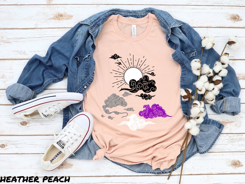 以下が含まれることがあります： ピーチ色のTシャツに、黒、グレー、白、紫の太陽と雲のグラフィックがプリントされています。雲は、ジェンダーニュートラルなプライドフラッグのようにスタイリッシュにデザインされています。