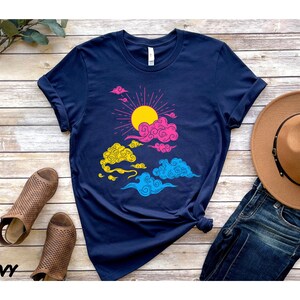 May include: A navy blue t-shirt with a graphic of a sun and clouds in pink, yellow, and blue. The clouds are stylized and have a whimsical look.