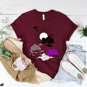 以下が含まれることがあります： 黒と白の太陽と雲のデザインが施されたマルーンのTシャツ。雲はグレー、パープル、ホワイトのグラデーション。太陽は白い中心に黒い光線がある。