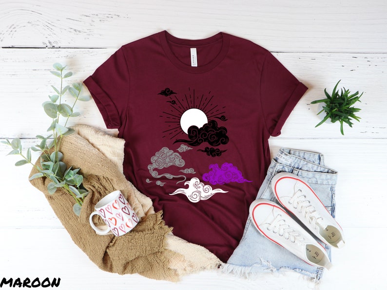 以下が含まれることがあります： 黒と白の太陽と雲のデザインが施されたマルーンのTシャツ。雲はグレー、パープル、ホワイトのグラデーション。太陽は白い中心に黒い光線がある。