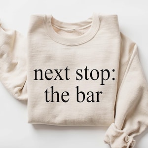 May include: A cream-coloured sweatshirt with the text "next stop: the bar" in black. The long sleeves are folded at the wrists. The sweatshirt is made of a soft material and is folded neatly.