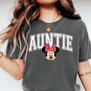 May include: Dark gray t-shirt with the word "AUNTIE" in white block letters and a Minnie Mouse graphic. The Minnie Mouse graphic features a red bow with white polka dots. The shirt is paired with blue jeans and white sneakers.