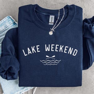 May include: A navy blue crewneck sweatshirt featuring the white text LAKE WEEKEND arched above a graphic of two crossed oars and stylized water waves. A silver chain necklace rests on the collar, and light blue denim jeans are visible underneath.