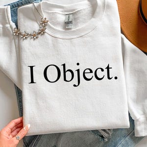 May include: A white sweatshirt with the text "I Object." in black, paired with blue jeans, a floral necklace, and a brown hat. The sweatshirt is a casual, everyday garment.
