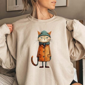 May include: Beige sweatshirt featuring a whimsical illustration of a cat wearing a teal beanie, scarf, and orange coat. The cat has a grumpy expression. The sweatshirt is a neutral color, suitable for casual wear.