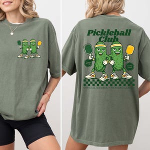 May include: A moss green t-shirt featuring a small graphic on the front left chest of two cartoon pickles holding pickleball paddles. The back displays a large graphic with the text Pickleball Club, two pickles, and the words Official Member Weekend League.