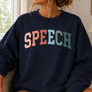 May include: A navy blue crewneck sweatshirt featuring the word SPEECH printed in a large, arched, collegiate-style font across the chest. The letters are colored in a gradient of soft peach, light blue, sage green, and dark teal.
