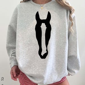 K&ouml;nnte beinhalten: Ein hellgraues Sweatshirt mit Rundhalsausschnitt und einer gro&szlig;en, einfarbig schwarzen Silhouette eines Pferdekopfes in der Mitte der Brust. Das Kleidungsst&uuml;ck hat lange &Auml;rmel und einen gerippten Ausschnitt f&uuml;r einen schlichten Look.
