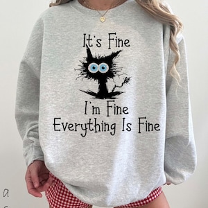 May include: A light gray crewneck sweatshirt featuring a black graphic of a messy-haired cat with large blue eyes. The text "It's Fine, I'm Fine, Everything Is Fine" is printed in black lettering above and below the cat illustration.