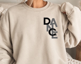 May include: A sand-colored crewneck sweatshirt featuring the word DANCE printed in a vertical, stacked layout on the upper left chest. The letters are rendered in a mix of solid black and grey, with a clean, modern sans-serif typeface design.