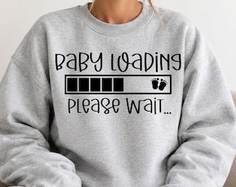 Puede incluir: Una sudadera gris claro de cuello redondo con texto negro que dice "BABY LOADING" sobre un gráfico de barra de carga rectangular con un pequeño par de huellas de bebé al final, seguido del texto "PLEASE WAIT..." en fuente cursiva.
