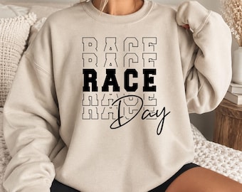 May include: A sand-colored crewneck sweatshirt featuring a graphic print on the chest. The design consists of the word RACE repeated in a stacked block font, with the middle instance in solid black, and the word Day in a black cursive script.