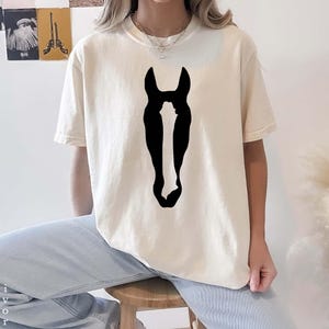 K&ouml;nnte beinhalten: Ein elfenbeinfarbenes Kurzarm-T-Shirt mit einer gro&szlig;en, einfarbig schwarzen Grafik einer Pferdekopf-Silhouette, die mittig auf der Brust platziert ist. Das Shirt verf&uuml;gt &uuml;ber einen klassischen Rundhalsausschnitt und eine lockere Passform.