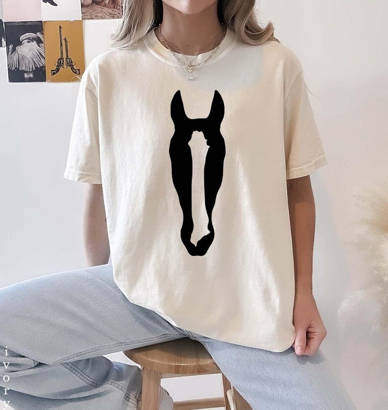 K&ouml;nnte beinhalten: Ein elfenbeinfarbenes Kurzarm-T-Shirt mit einer gro&szlig;en, einfarbig schwarzen Grafik einer Pferdekopf-Silhouette, die mittig auf der Brust platziert ist. Das Shirt verf&uuml;gt &uuml;ber einen klassischen Rundhalsausschnitt und eine lockere Passform.