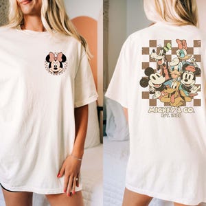 Op de afbeelding: Cr&egrave;mekleurig T-shirt met een Minnie Mouse-afbeelding op de voorkant en een groep Disney-personages op de achterkant. De afbeelding op de achterkant bevat Mickey Mouse, Donald Duck, Goofy en Daisy Duck. Het shirt heeft korte mouwen en een relaxte pasvorm.