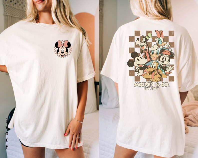 Op de afbeelding: Cr&egrave;mekleurig T-shirt met een Minnie Mouse-afbeelding op de voorkant en een groep Disney-personages op de achterkant. De afbeelding op de achterkant bevat Mickey Mouse, Donald Duck, Goofy en Daisy Duck. Het shirt heeft korte mouwen en een relaxte pasvorm.