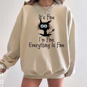 May include: A beige crewneck sweatshirt featuring a black graphic of a disheveled cat with large blue eyes. The text "It's Fine I'm Fine Everything Is Fine" is printed in black lettering above and below the cat illustration on the front.