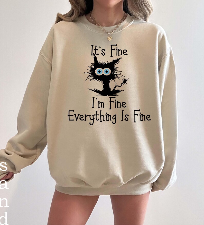 May include: A beige crewneck sweatshirt featuring a black graphic of a disheveled cat with large blue eyes. The text "It's Fine I'm Fine Everything Is Fine" is printed in black lettering above and below the cat illustration on the front.