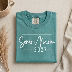 Może przedstawiać: Turkusowy t-shirt z kr&oacute;tkim rękawem z białym prostokątnym obrysem i nadrukowanym wewnątrz napisem Senior Mom 2027. Koszulka ma okrągły dekolt oraz widoczną metkę marki Comfort Colors wewnątrz przy kołnierzyku.