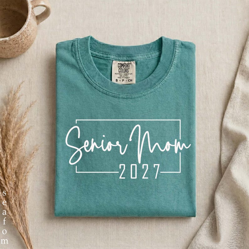 Może przedstawiać: Turkusowy t-shirt z kr&oacute;tkim rękawem z białym prostokątnym obrysem i nadrukowanym wewnątrz napisem Senior Mom 2027. Koszulka ma okrągły dekolt oraz widoczną metkę marki Comfort Colors wewnątrz przy kołnierzyku.