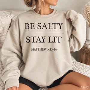 May include: A sand-colored crewneck sweatshirt featuring black text that reads BE SALTY STAY LIT with the reference MATTHEW 5:13-14 printed below. The garment has long sleeves, a ribbed neckline, and a standard fit, showcasing a neutral tone.