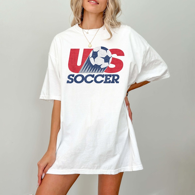 Pu&ograve; includere: Una t-shirt bianca a maniche corte con un design grafico sul petto. La grafica mostra le lettere rosse U e S con un pallone da calcio bianco e nero che sostituisce la lettera O, seguita dalla parola SOCCER in lettere maiuscole blu.