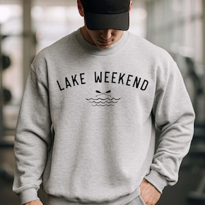 May include: A light gray crewneck sweatshirt featuring the black text LAKE WEEKEND and a graphic of two crossed oars above wavy lines, paired with a matching light gray baseball cap. The sweatshirt has long sleeves and a ribbed hem and cuffs.