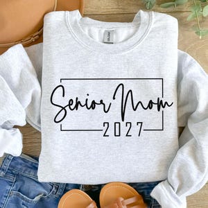 Może przedstawiać: Jasnoszara bluza z okrągłym dekoltem z czarnym napisem Senior Mom 2027 umieszczonym w cienkiej czarnej prostokątnej ramce. Odzież posiada długie rękawy z prążkowanymi mankietami oraz prążkowany dekolt, co nadaje jej prosty wygląd.
