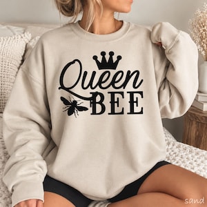 May include: A sand-colored crewneck sweatshirt featuring a black graphic print on the chest. The design includes a stylized crown above the word Queen in cursive script, followed by the word BEE in block letters and a small bee illustration.