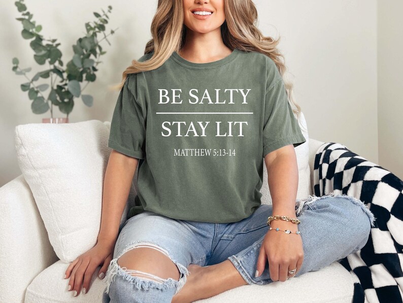 May include: Olive green t-shirt with the text "BE SALTY STAY LIT" and "MATTHEW 5:13-14" in white. The shirt is worn with light blue jeans. The overall theme is casual wear.